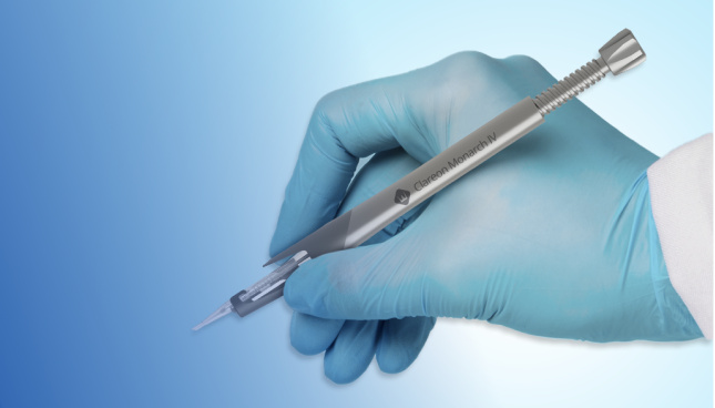 Ein Bild des Clareon Monarch IV Implantationssystems wie ein Stift gehalten von einer Hand in einem blauen chirurgischen Handschuh vor blauem Hintergrund.
