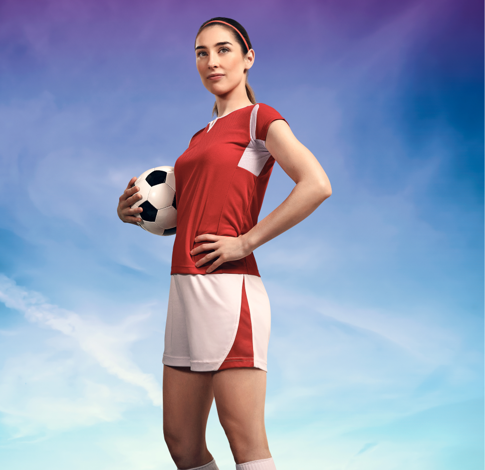 Jugadora de fútbol con uniforme rojo y blanco sosteniendo un balón, posando con confianza frente a un cielo azul y púrpura