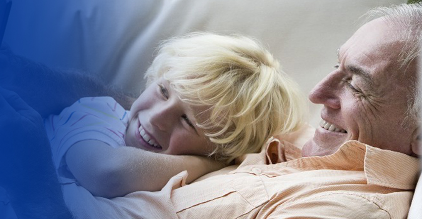 An image of an older man and his grandson sitting on a couch smiling and looking at a tablet that appears off-screen.