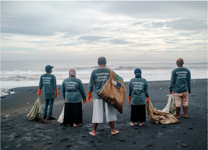 A group of men and women at the beach wearing shirts that say “Stopping Ocean Plastic” while holding bags of collected rubbish