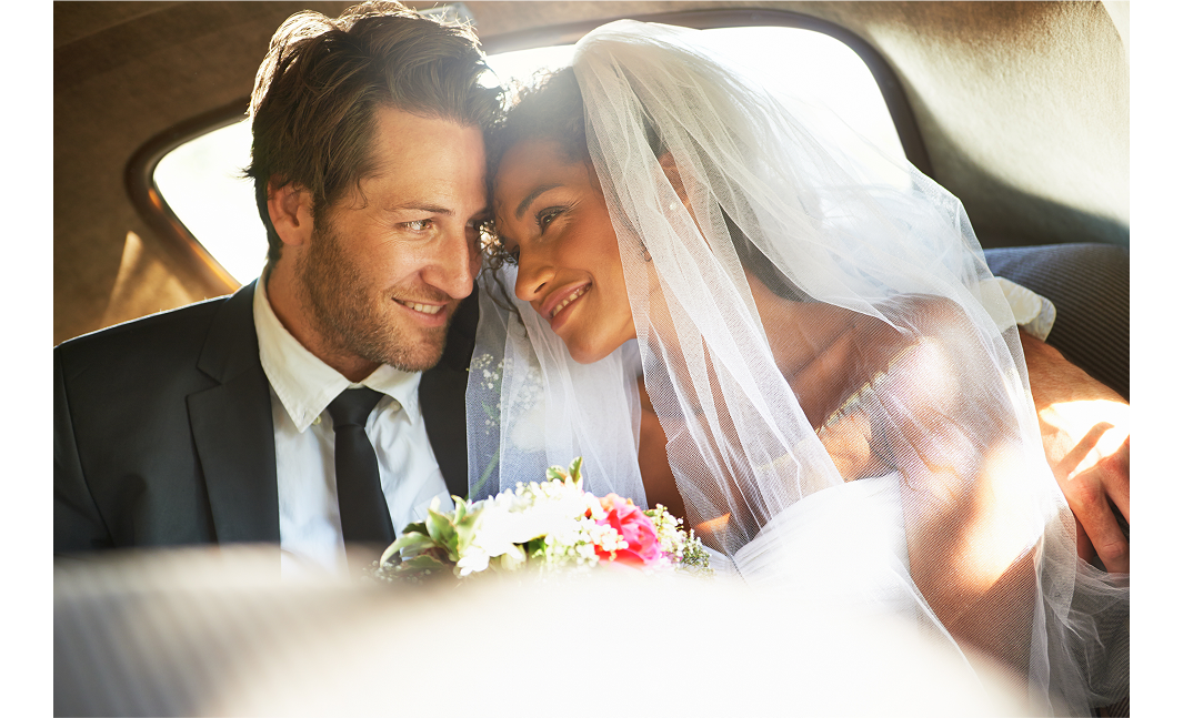 A married couple sitting side by side in a car. They are touching foreheads while smiling at each other. The man is wearing a black suit, with a white shirt and black tie. The woman is wearing a white wedding dress. A bouquet of white and pink flowers appears between them.