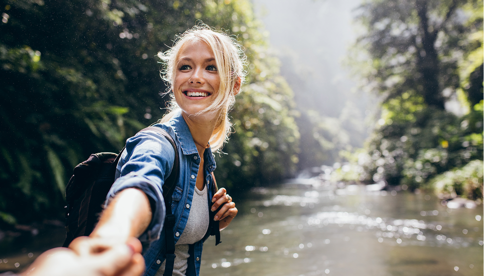 A smiling woman taken from the perspective of the person holding her hand. She is wearing a black backpack and a blue collared shirt over a white t-shirt. Green trees and a dark-colored river appear in the background.
