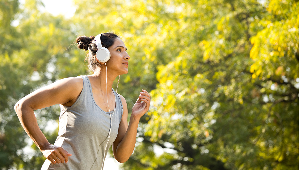 A woman running in a gray tank top with white over-the-ear headphones. Green trees appear in the background.