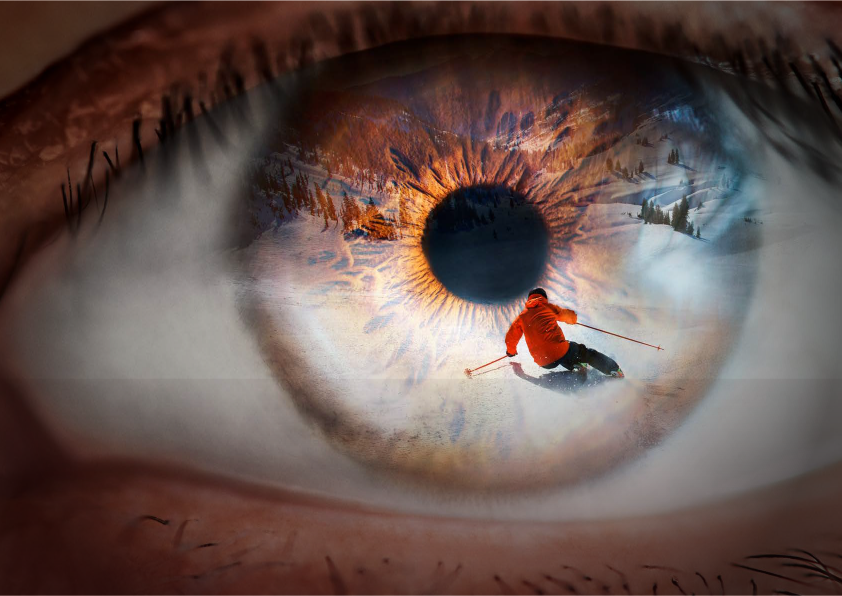 Close-up of a human eye, in the iris of which a skier in a red jacket riding down a snowy slope is reflected.