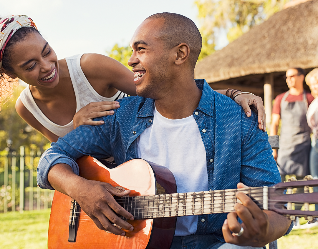 A smiling man in a collared blue shirt over a white t-shirt is playing guitar outside. A smiling woman in a white tank top appears over his shoulder. Both are smiling at each other.