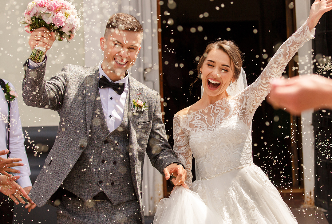 A smiling man and woman on their wedding day. The man is in a gray suit carrying a pink bouquet of flowers and holding hands with the woman who is in a white dress with her arm in the arm. Confetti appears around them.