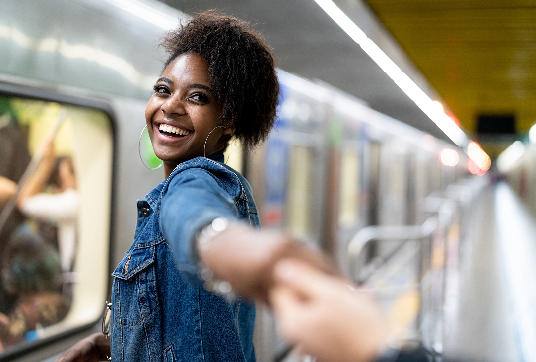 A smiling woman looks back as she holds a man's hand and they walk together through a busy subway station.