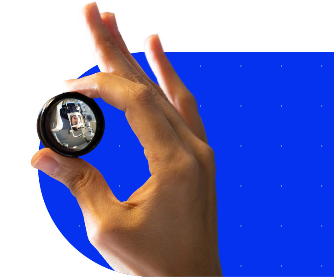 An image of a hand holding up a lens. Through the lens, we see an inverted image of a patient during a slit lamp examination.