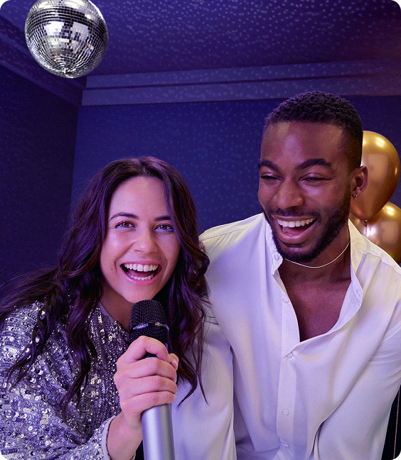 Woman holding a microphone and smiling next to a man, both laughing under a disco ball at a party with balloons in the background