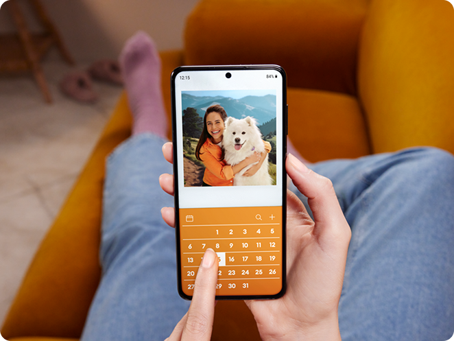 Person holding a smartphone while sitting on a couch, viewing a photo of a woman hugging a fluffy white dog, with a calendar interface below the image