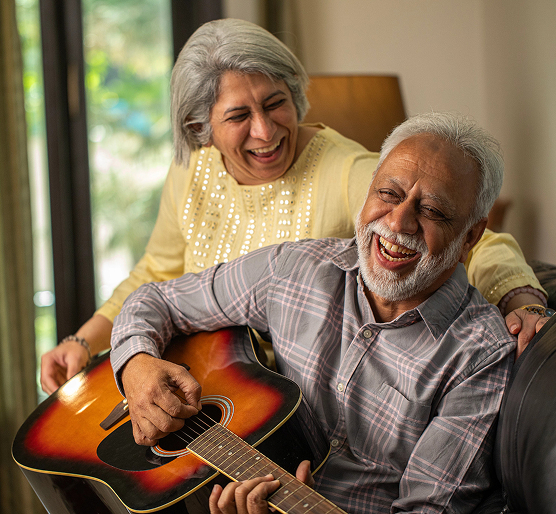 Couple âgé riant ensemble à la maison, l’homme jouant de la guitare, illustrant des activités quotidiennes agréables et la qualité de vie.