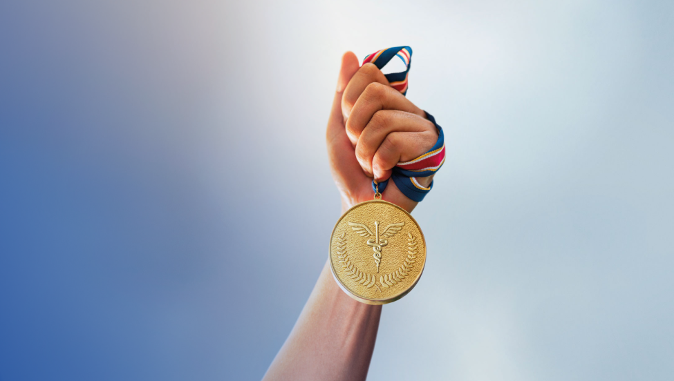An image of an arm in the foreground with a blue and white background, featuring a hand holding an intertwined gold medal with a red and blue ribbon.