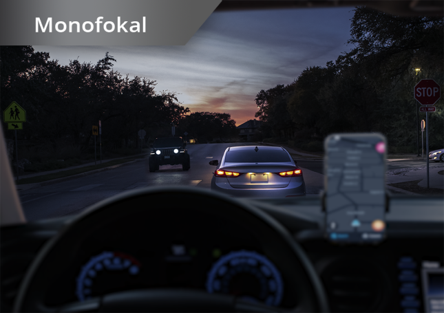 Blick aus einem Auto heraus auf eine Straße in der Dämmerung. Ein Smartphone ist auf dem Armaturenbrett befestigt. Der weiße Text links oben besagt “Monofokallinse”.