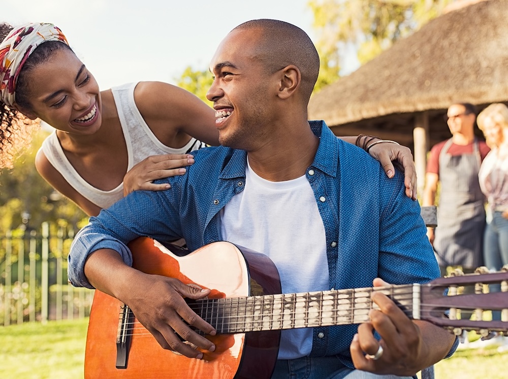 Mann sitzt und spielt Gitarre, während eine Frau lächelt und bei einer Outdoor-Veranstaltung eine Hand auf seine Schulter legt