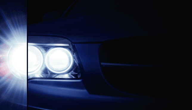 Nahaufnahme eines linken Scheinwerfers eines Autos, der die Dunkelheit erhellt. Eine vertikale Linie teilt den Scheinwerfer in zwei Hälften, wobei das Licht auf der linken Hälfte einen Starburst-Effekt aufweist.
