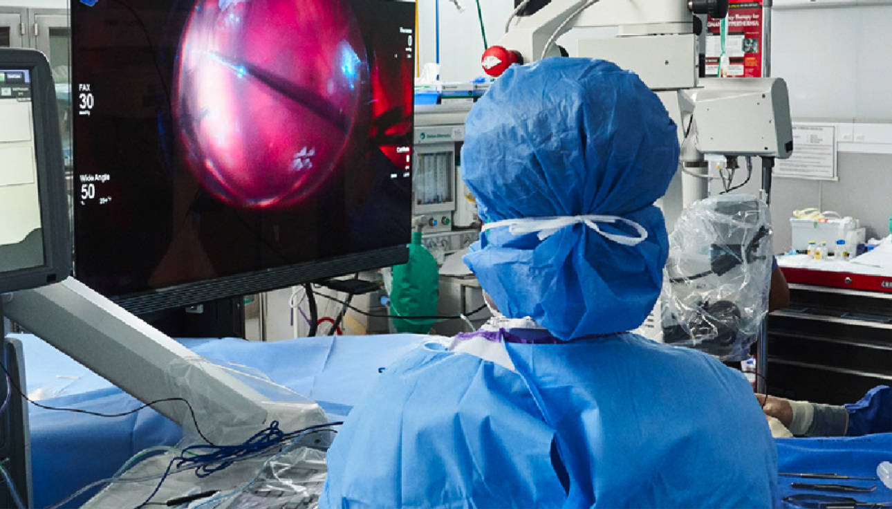 Das Bild einer Gruppe Chirurgen im OP-Saal mit einem Patienten, der operiert wird. Ein NGENUITY Bildschirm zeigt eine Vergrößerung der Operation für die Anwesenden im Raum.