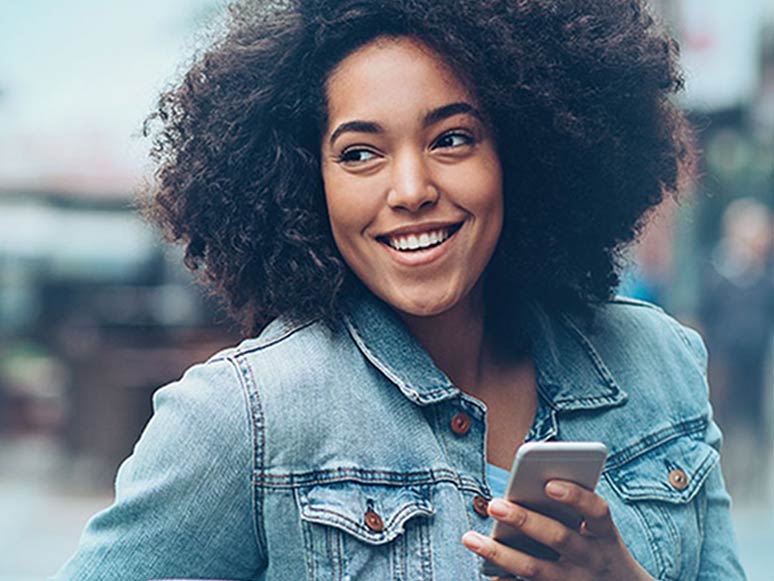 Lächelnde Frau in Jeansjacke, die draußen sitzt, mit einer Kaffeetasse in der einen und einem Telefon in der anderen Hand