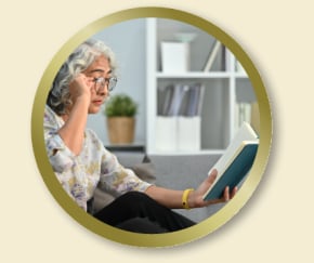 Personne portant des lunettes, assise sur un canapé, lisant un livre dans un salon lumineux avec une bibliothèque en arrière-plan.
