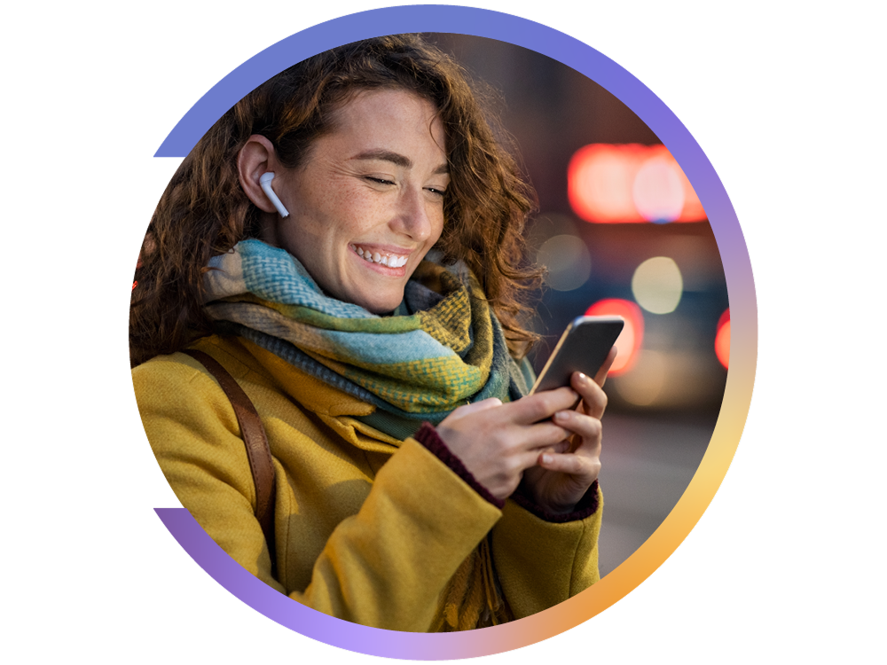 Femme souriante avec manteau jaune et foulard, regardant son téléphone tout en portant des écouteurs sans fil