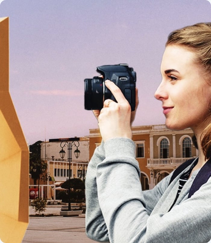 Personne tenant un appareil photo et photographiant une place urbaine bordée de bâtiments sous un ciel aux teintes douces.