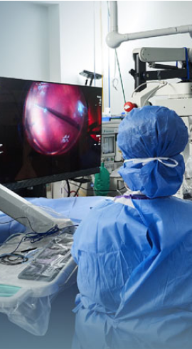 Εικόνα ενός χειρουργού ο οποίος φοράει μπλε χειρουργική ποδιά στο χειρουργείο και παρακολουθεί μια χειρουργική επέμβαση στην οθόνη.