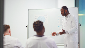 An image of a group people wearing lab coats while talking to each other, in front of a whiteboard