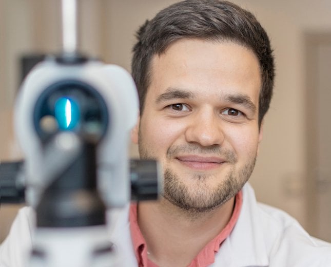 Close-up view of an ophthalmology diagnostic device with a bright blue light in the lens, positioned in front of a healthcare professional wearing a white coat.