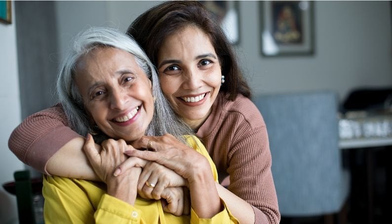 Two women sitting indoors, one wearing a mustard yellow top and the other in a long-sleeve brown top, embracing closely with arms wrapped around each other in a warm, supportive gesture. Background includes a blurred chair and framed artwork on the wall.