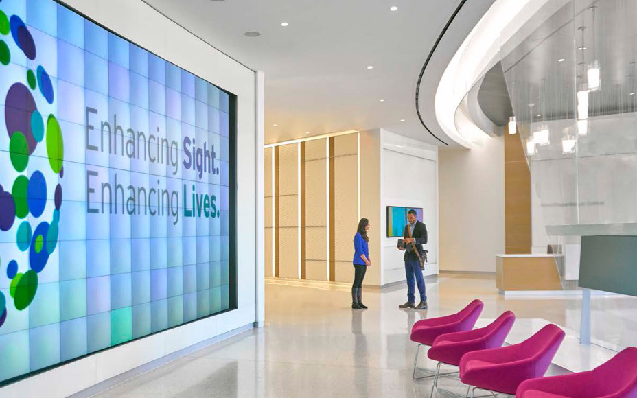Lobby at Alcon Experience Academy featuring a large digital display with the words 'Enhancing Sight. Enhancing Lives.', two people conversing near the center, and a row of magenta chairs in the foreground