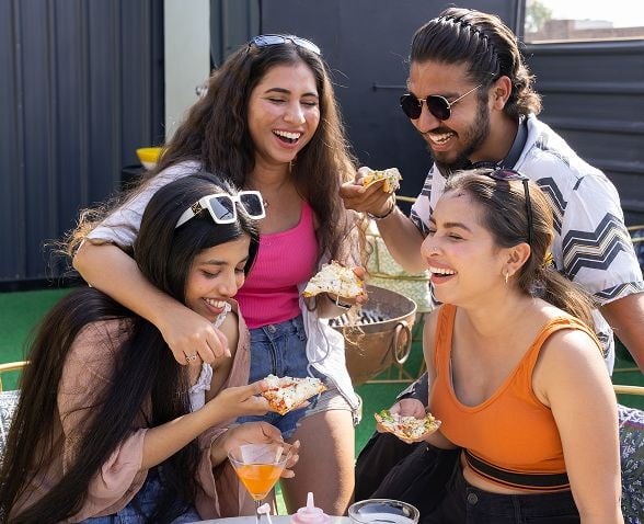 Group of friends laughing and sharing snacks outdoors, holding drinks and small plates, enjoying a casual social gathering in the sunshine.