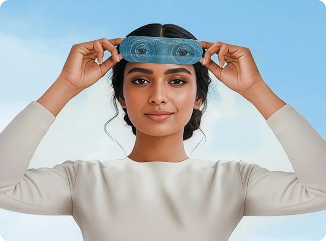 Person holding a blue, transparent eye mask with circular lens-like graphics against their forehead, looking at the camera against a light blue background.