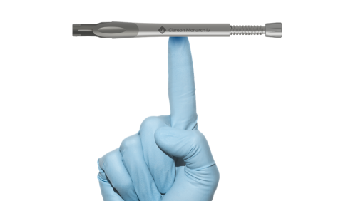 Hand wearing a disposable glove with the index finger pointing upwards. The Clareon Monarch IV Handpiece sits horizontally, balanced on top of the index finger.