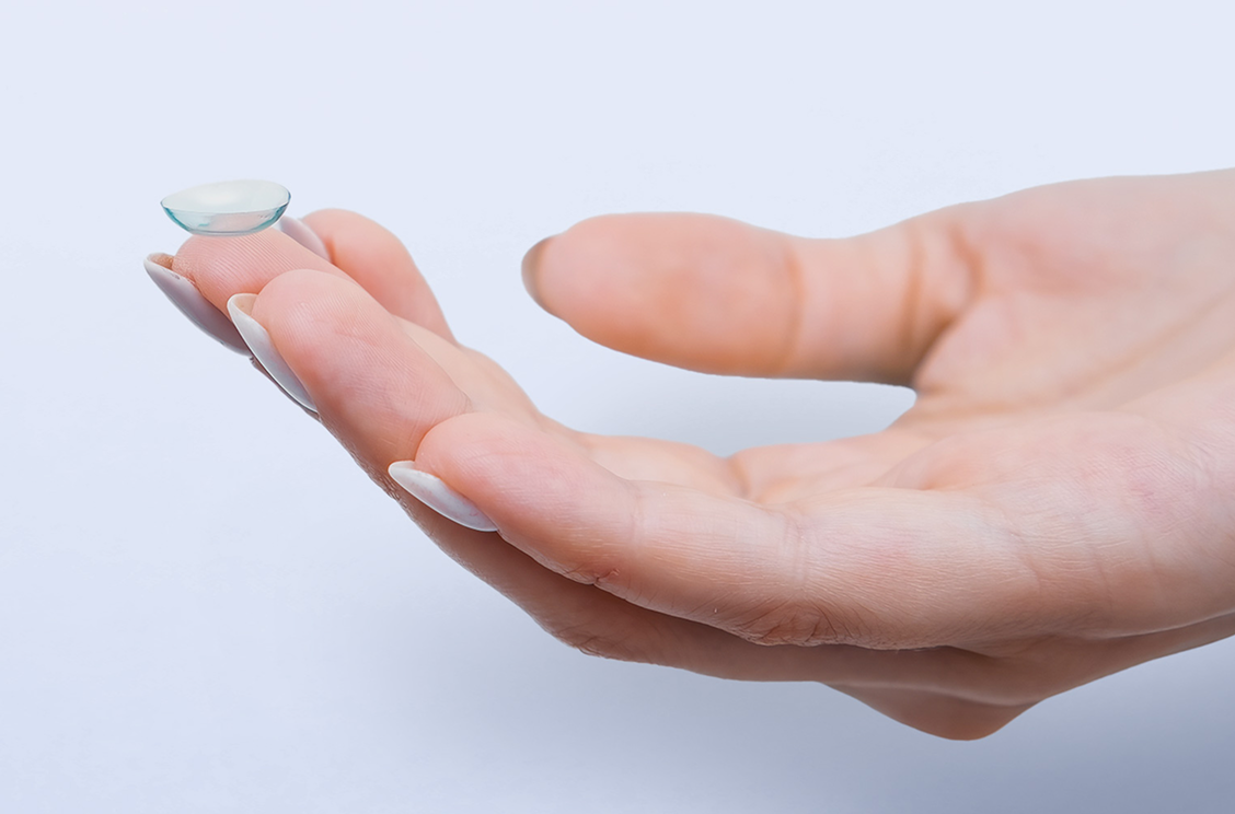 Close-up of a hand holding a contact lens on the fingertip, set against a gray background