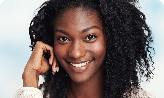 Confident woman with curly hair smiling warmly, resting her hand against her face in a soft-focus indoor setting