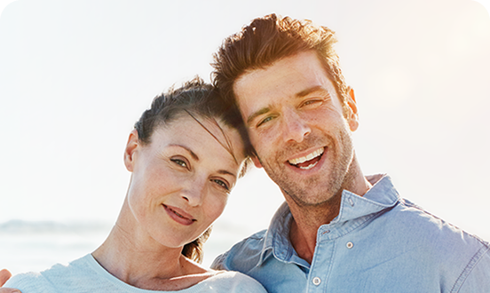 Smiling couple standing close together outdoors on a sunny day, with the ocean in the background.