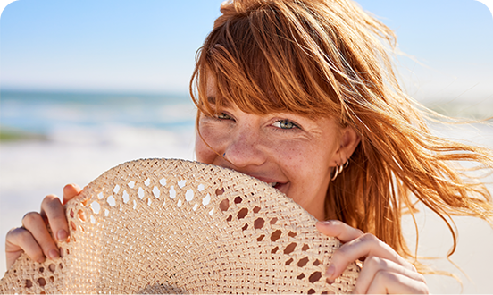 Smiling woman with red hair holding a woven sun hat in front of her face on a sunny beach