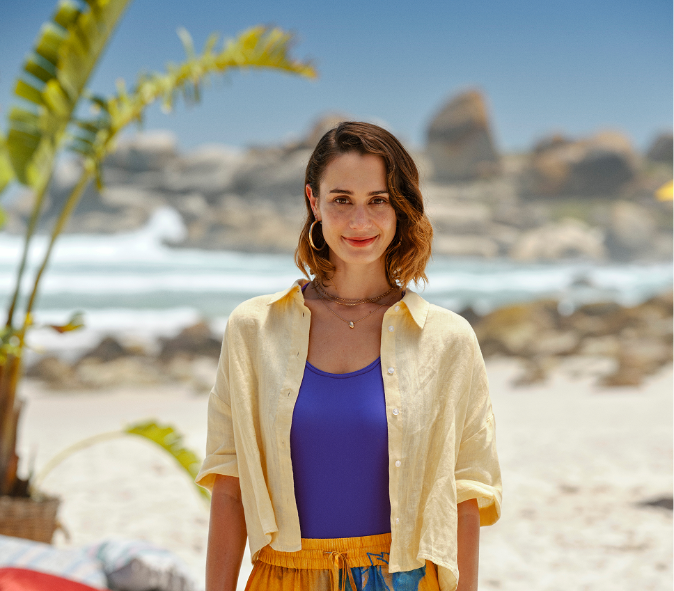 Mujer joven sonriente con blusa amarilla, traje de baño morado y pantalones coloridos, de pie en una playa soleada con palmeras y rocas al fondo.