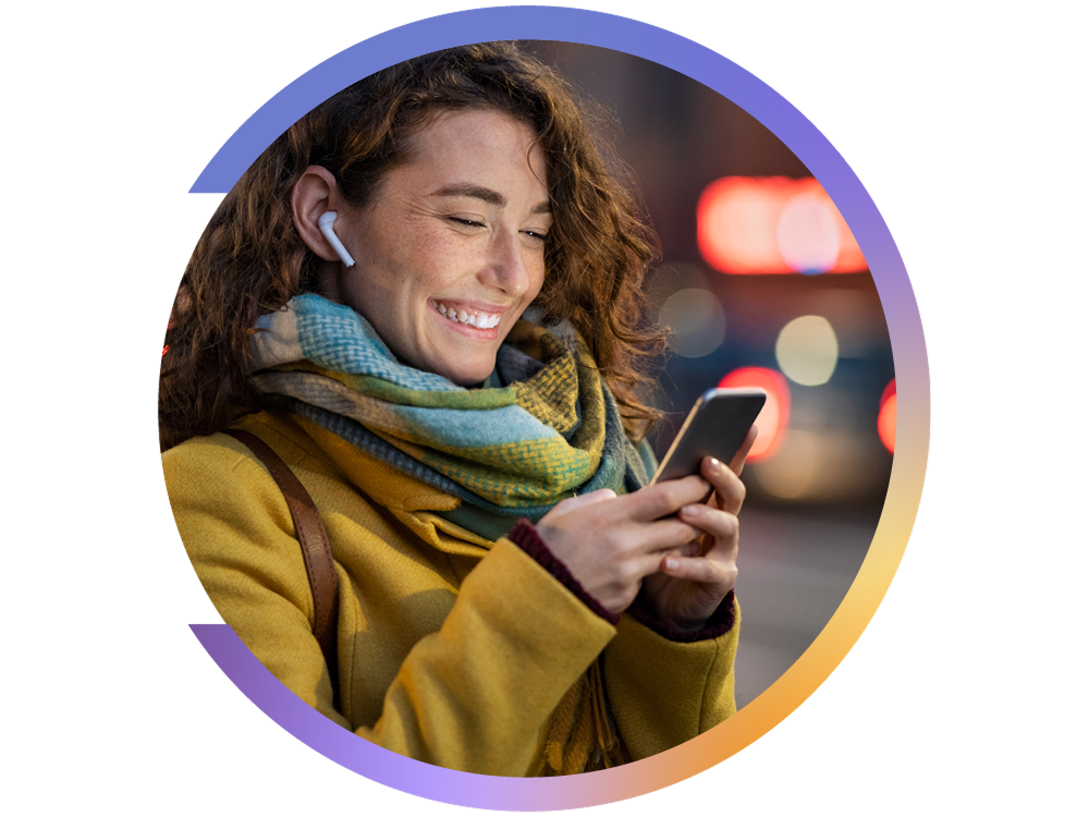 Mulher sorridente com casaco amarelo e cachecol, a olhar para o telemóvel enquanto usa auriculares sem fios