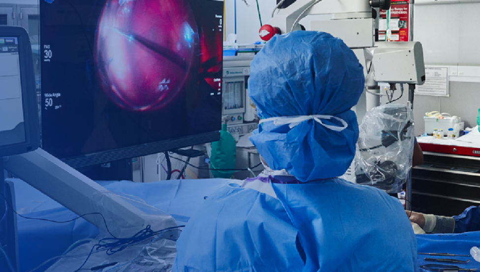 An image of a group of surgeons in an operating room with a patient undergoing surgery. An NGENUITY screen shows a close-up view of the surgery to the surgeons in the room.