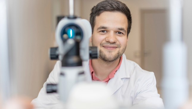 Friendly looking male optometrist standing behind eye exam equipment used to fit contact lenses