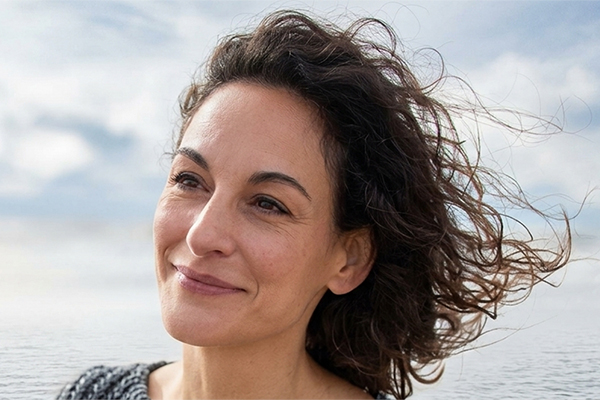 A woman with windblown hair standing near a calm ocean, looking toward the horizon under a bright, lightly clouded sky.