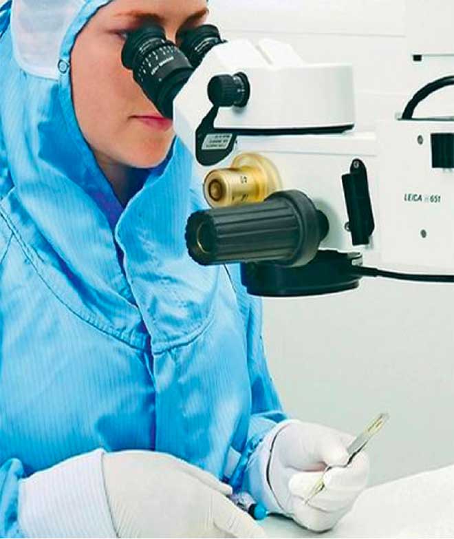 Person wearing a blue sterile gown and gloves performing a precise task with surgical instruments under a Leica microscope in a clean clinical environment.