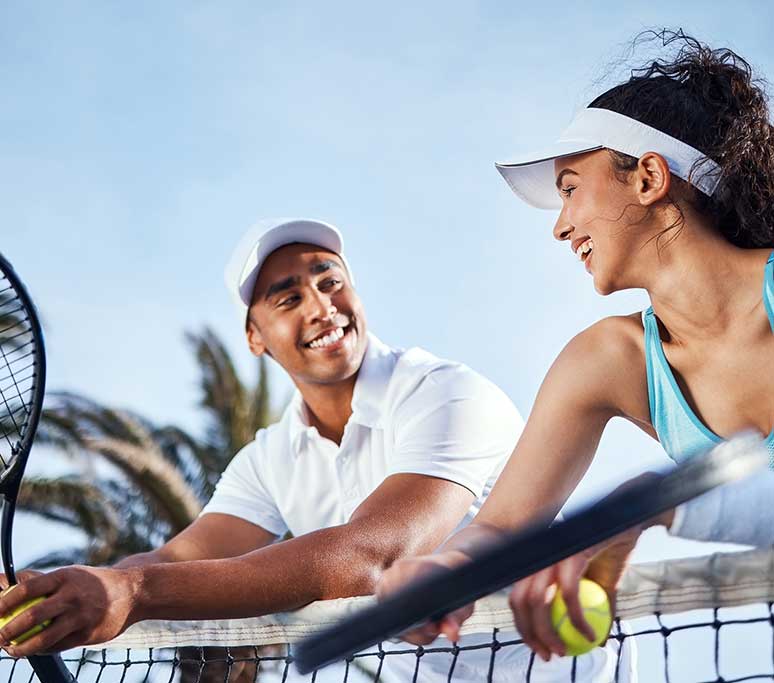 A man and woman smile at each other while holding tennis rackets outdoors
