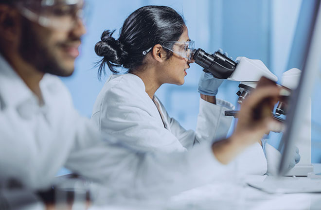 Laboratory setting with individuals wearing white lab coats working at microscopes and computer monitors. One person is closely observing a sample through a microscope while another is positioned near a computer screen. The background shows a clean, clinical environment with soft blue tones.