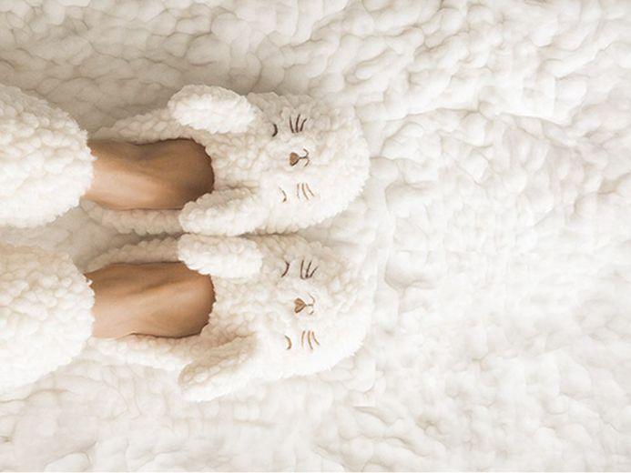 Person wearing fluffy white slippers shaped like sleepy animal faces with ears, resting on a soft, textured white blanket