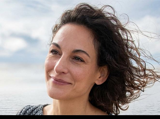 A woman with windblown hair standing near a calm ocean, looking toward the horizon under a bright, lightly clouded sky.