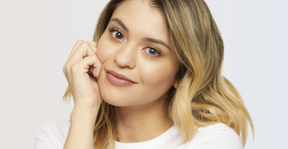 Smiling woman with blonde hair resting her chin on her hand, wearing a white top and one blue and one brown colored contact lens to show a side-by-side comparison effect.