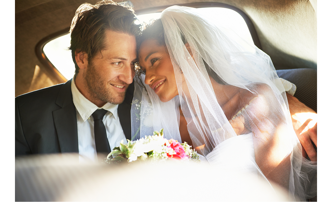 A married couple sitting side by side in a car. They are touching foreheads while smiling at each other. The man is wearing a black suit, with a white shirt and black tie. The woman is wearing a white wedding dress. A bouquet of white and pink flowers appears between them.