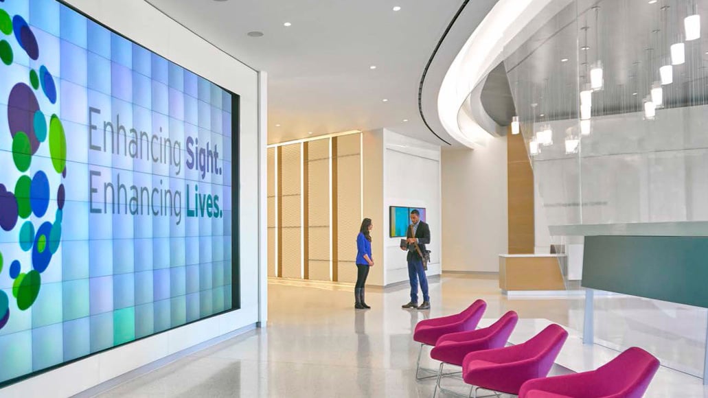 Modern Alcon office lobby with a large digital display reading “Enhancing Sight. Enhancing Lives.” and two professionals talking nearby.