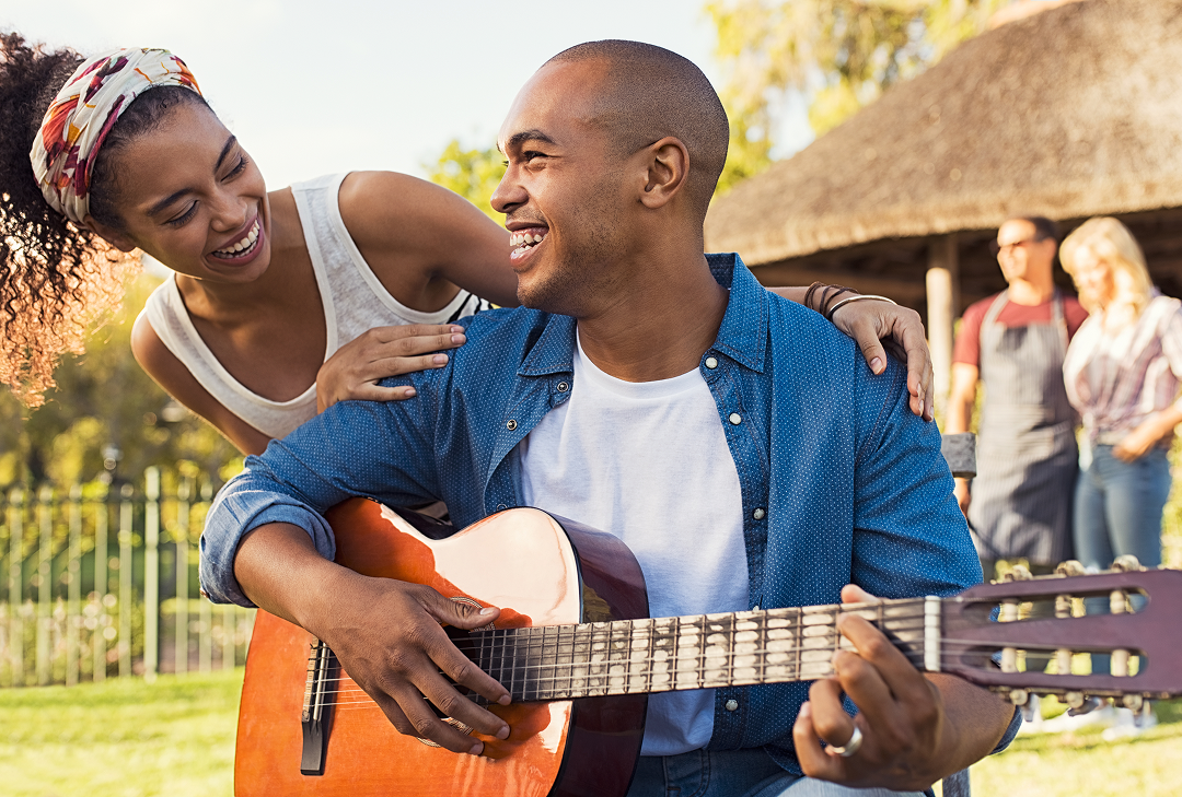 A smiling man in a collared blue shirt over a white t-shirt is playing guitar outside. A smiling woman in a white tank top appears over his shoulder. Both are smiling at each other.